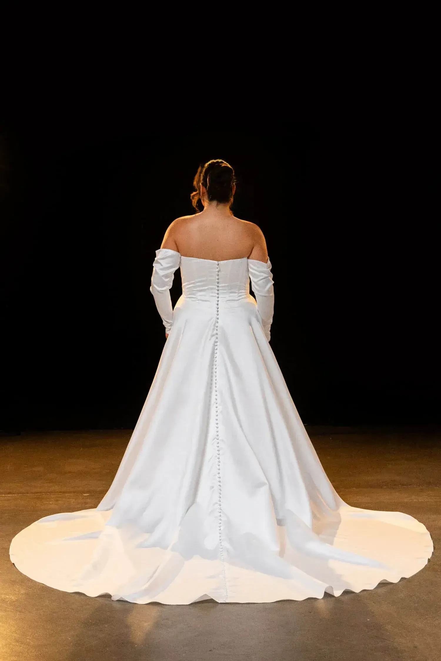 A woman in an elegant white wedding dress stands with her back facing the camera. The gown features off-the-shoulder sleeves and a long train, creating a graceful silhouette against a dark background.