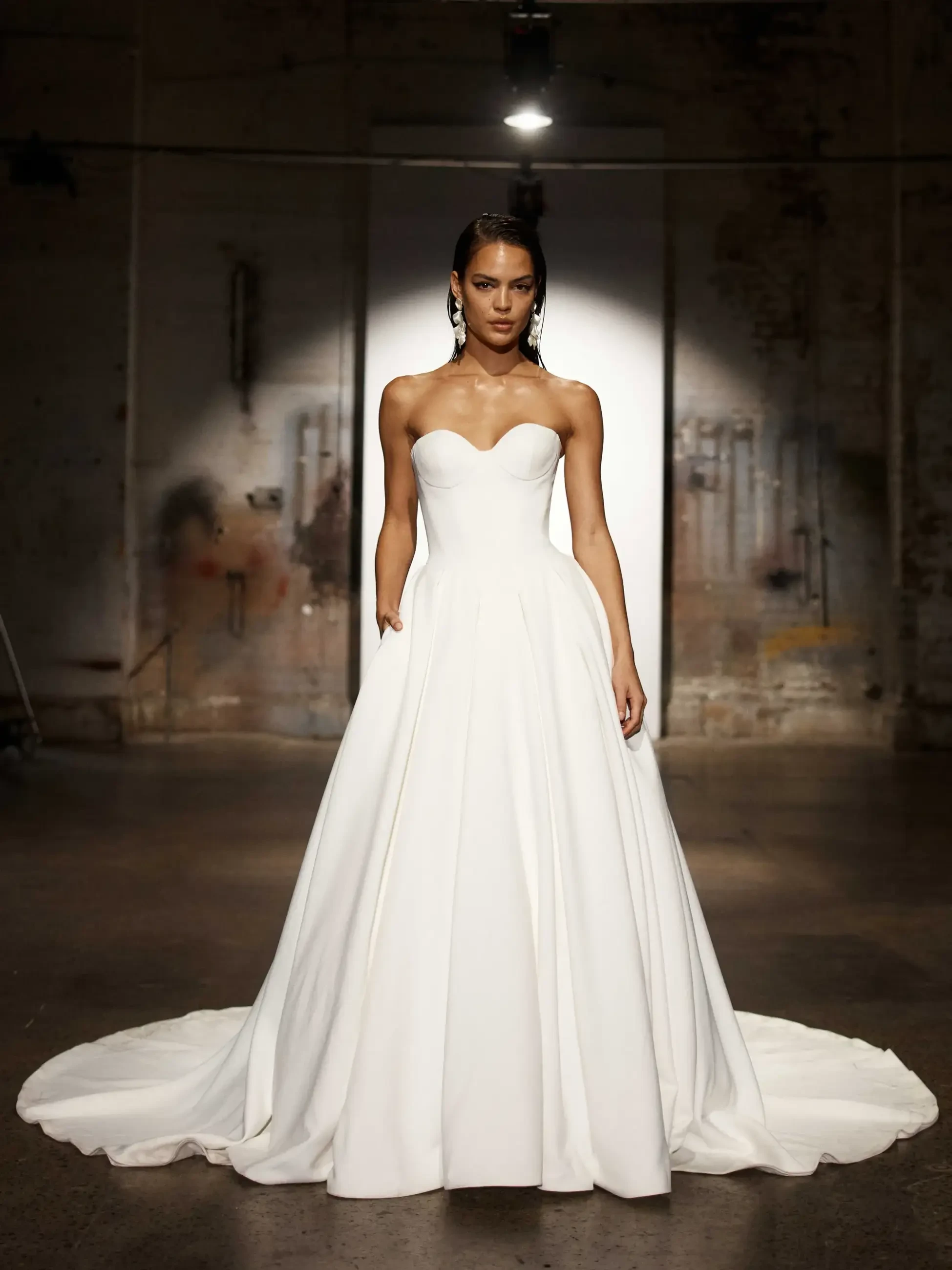 A model in a classic white strapless ball gown stands confidently under a spotlight in an industrial setting, with a serene and elegant expression.