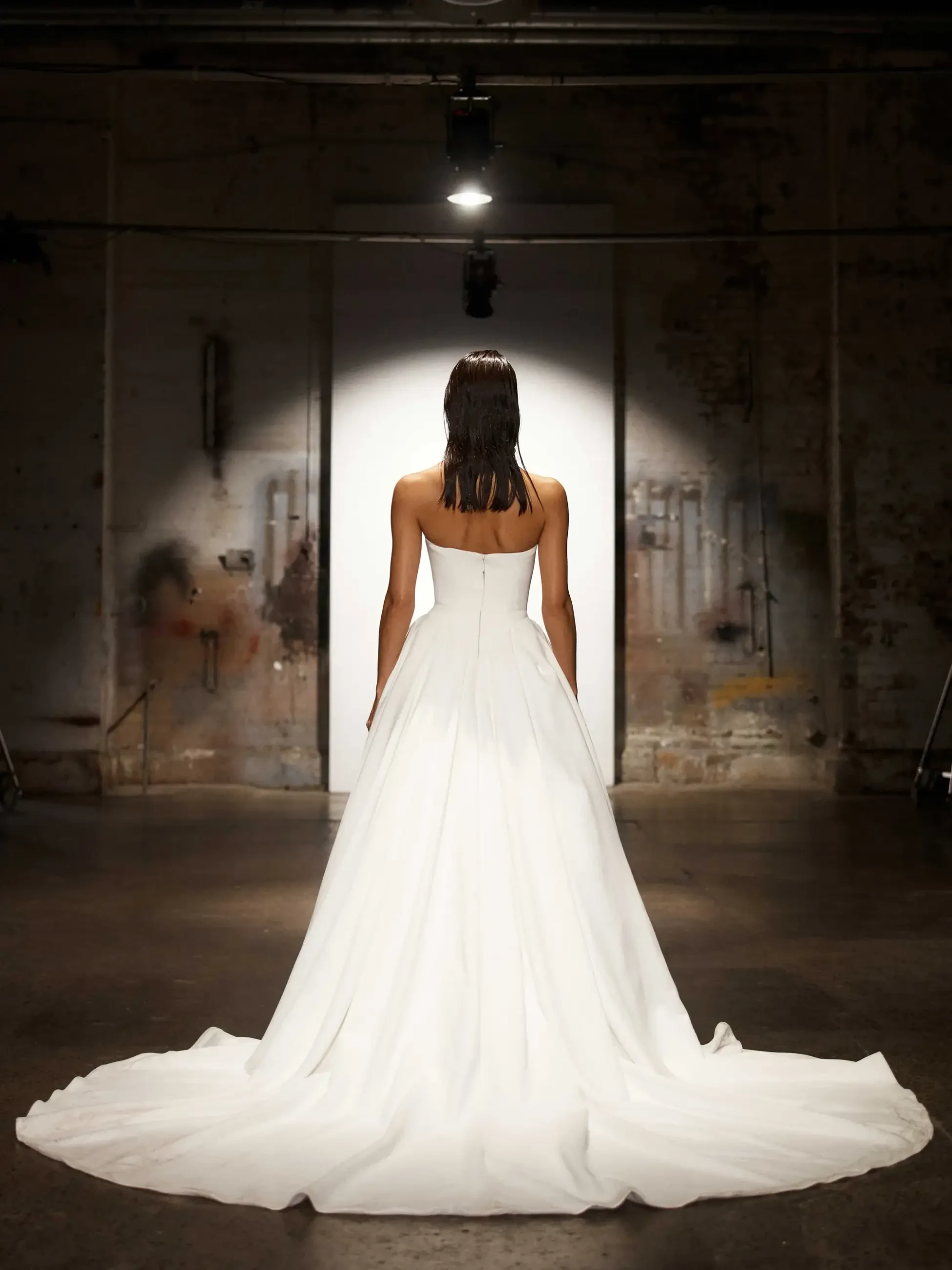 A woman in a long, elegant white gown stands facing a lit doorway in a dim, industrial setting. The mood is dramatic and focused.