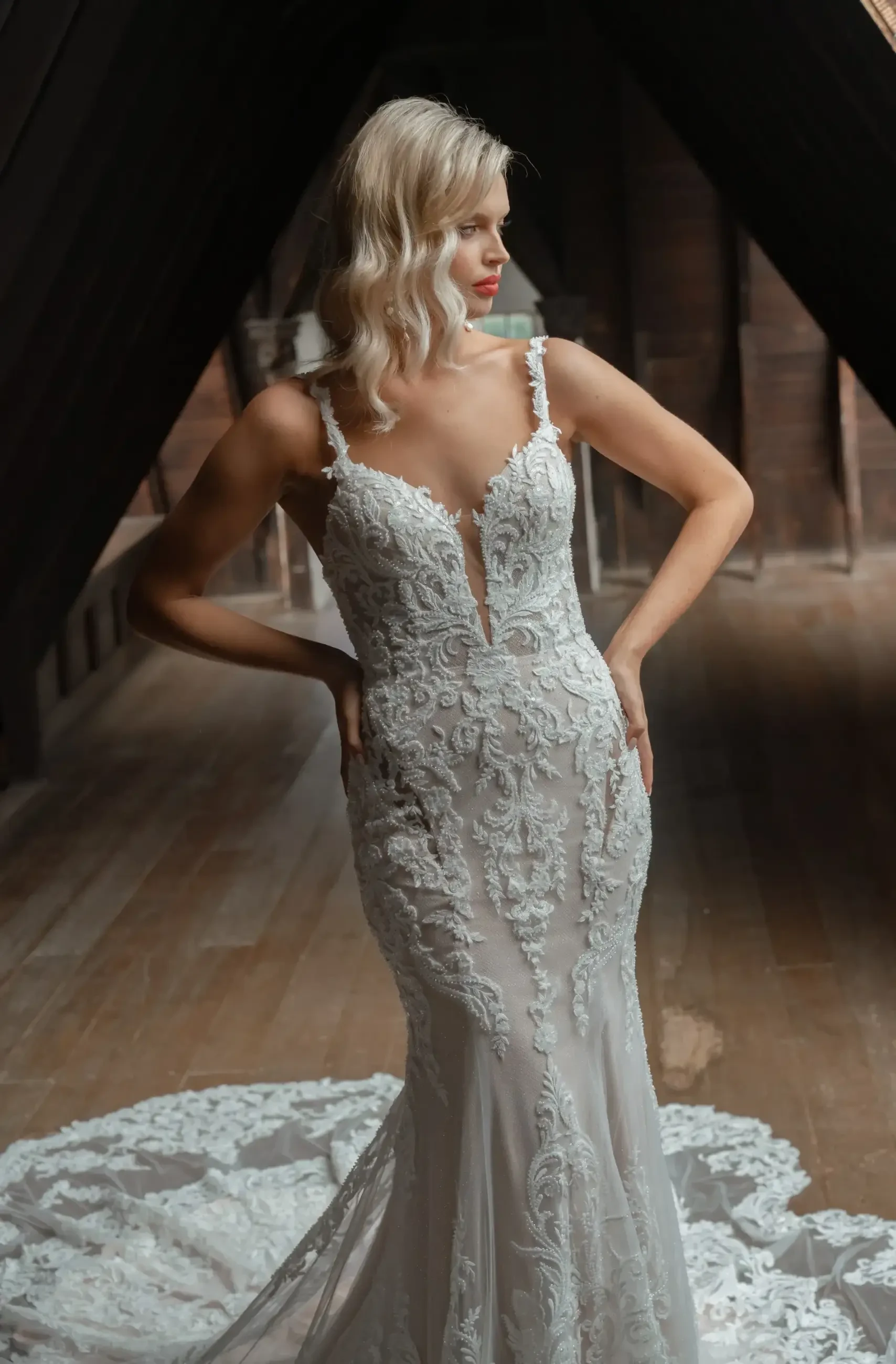 A bride in a fitted lace gown stands confidently in an elegant, dimly lit wooden hallway. Her wavy blonde hair complements the gown's intricate details.