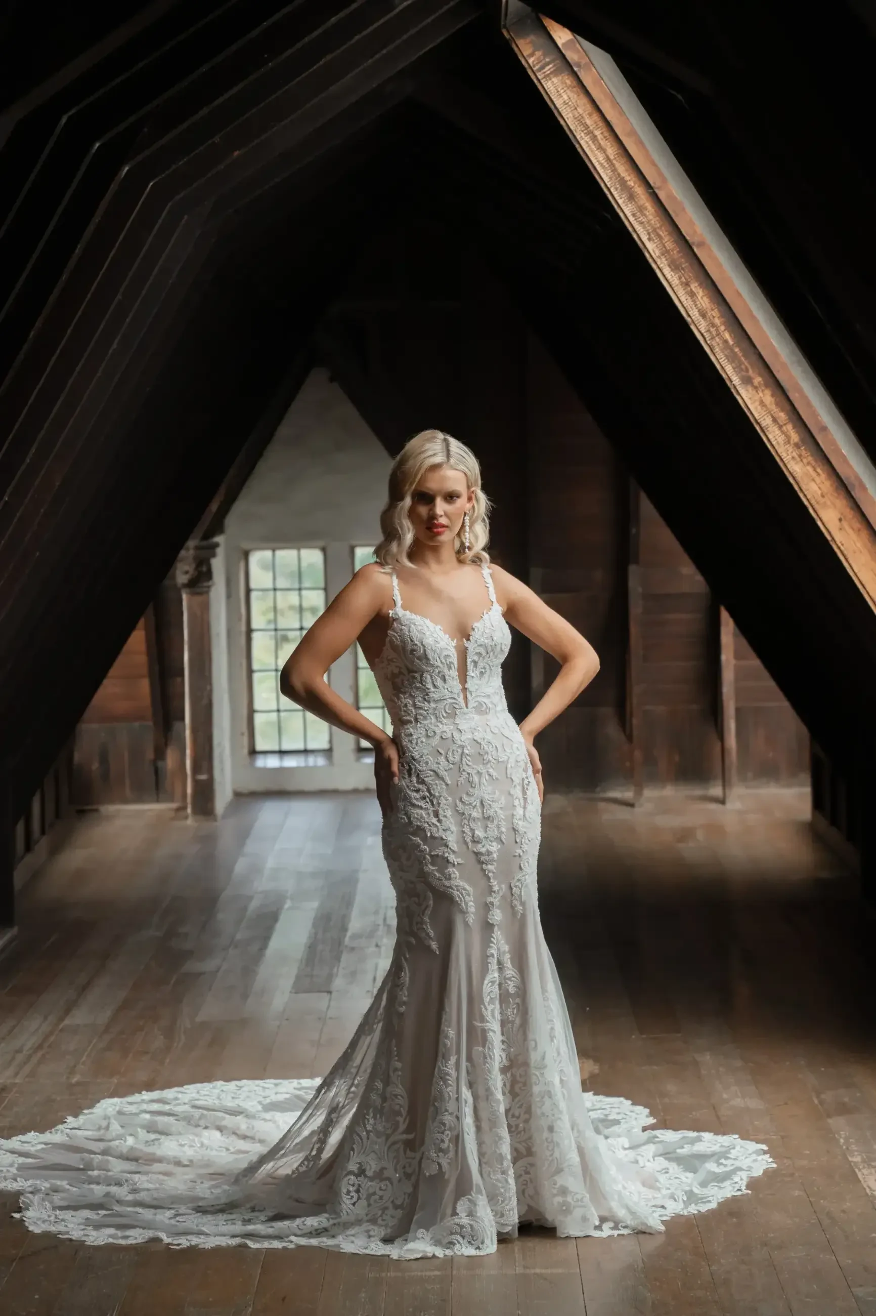 Bride stands confidently in an ornate lace gown with a long train, framed by a dramatic dark wooden archway, conveying elegance and poise.
