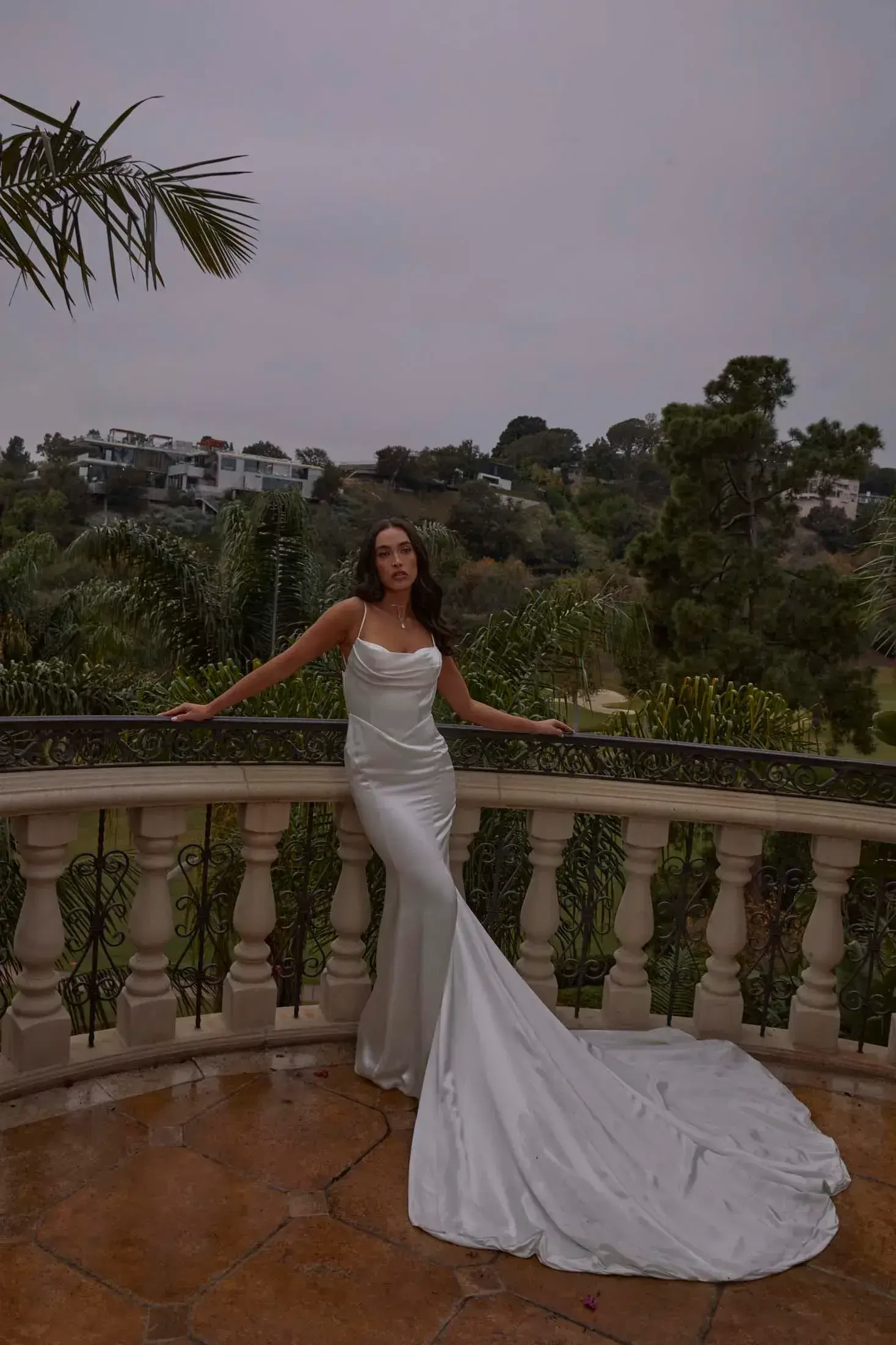 A woman in an elegant white gown with a long train stands confidently on a balcony, surrounded by greenery with distant hills under an overcast sky.