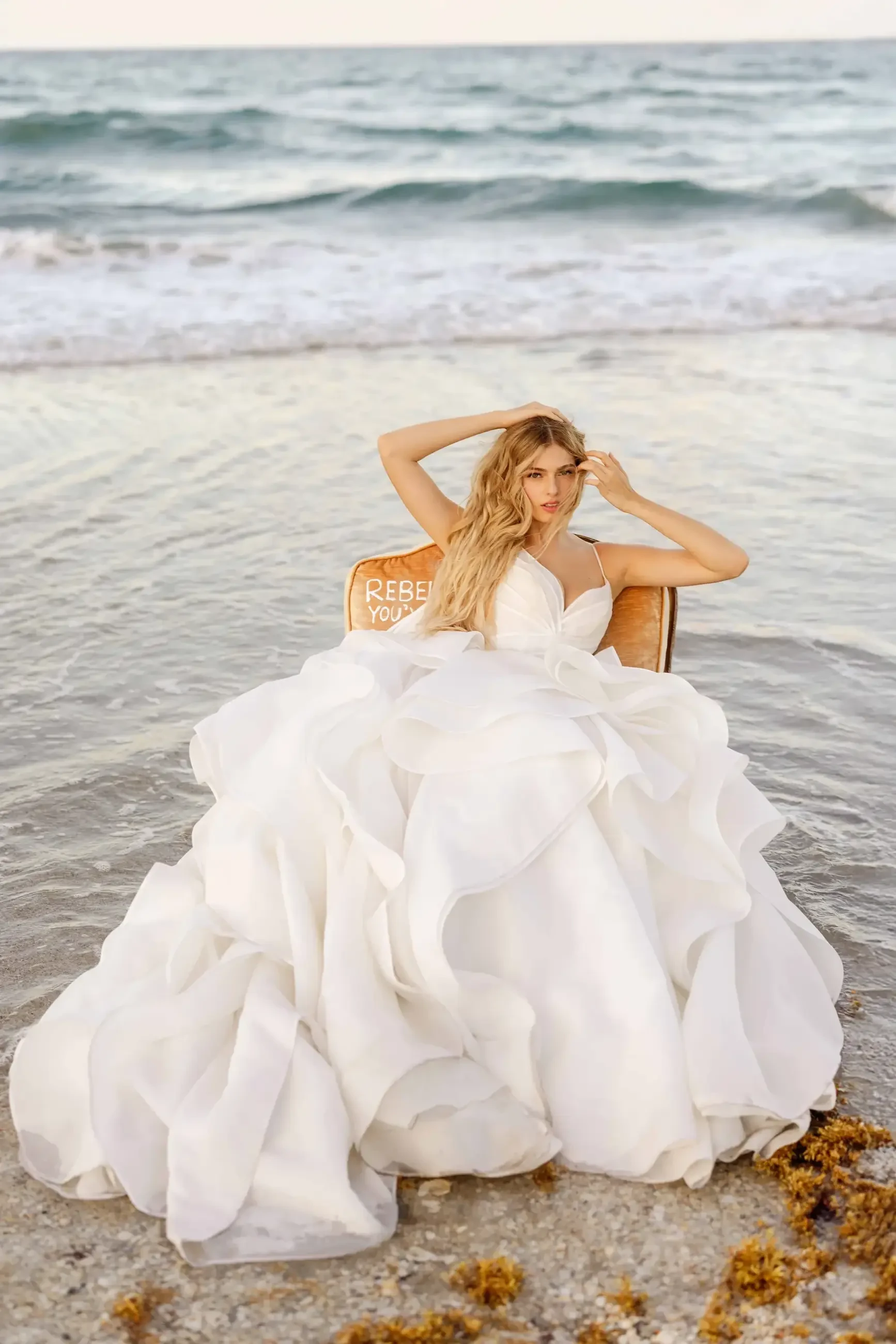 A person in a white gown sitting on a chair by the water with waves in the background.