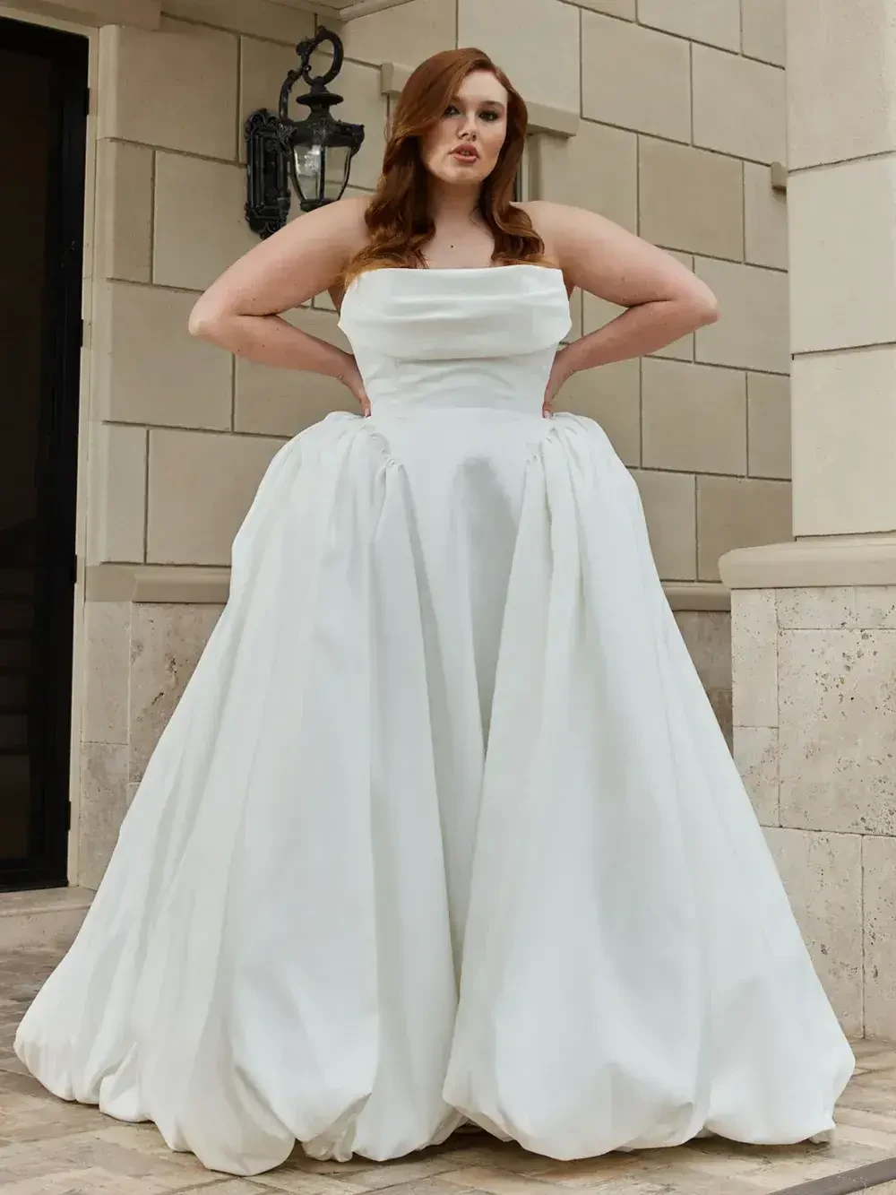 A woman wearing a strapless, white ball gown stands outside, with her hands on her hips, in front of a stone wall and door.