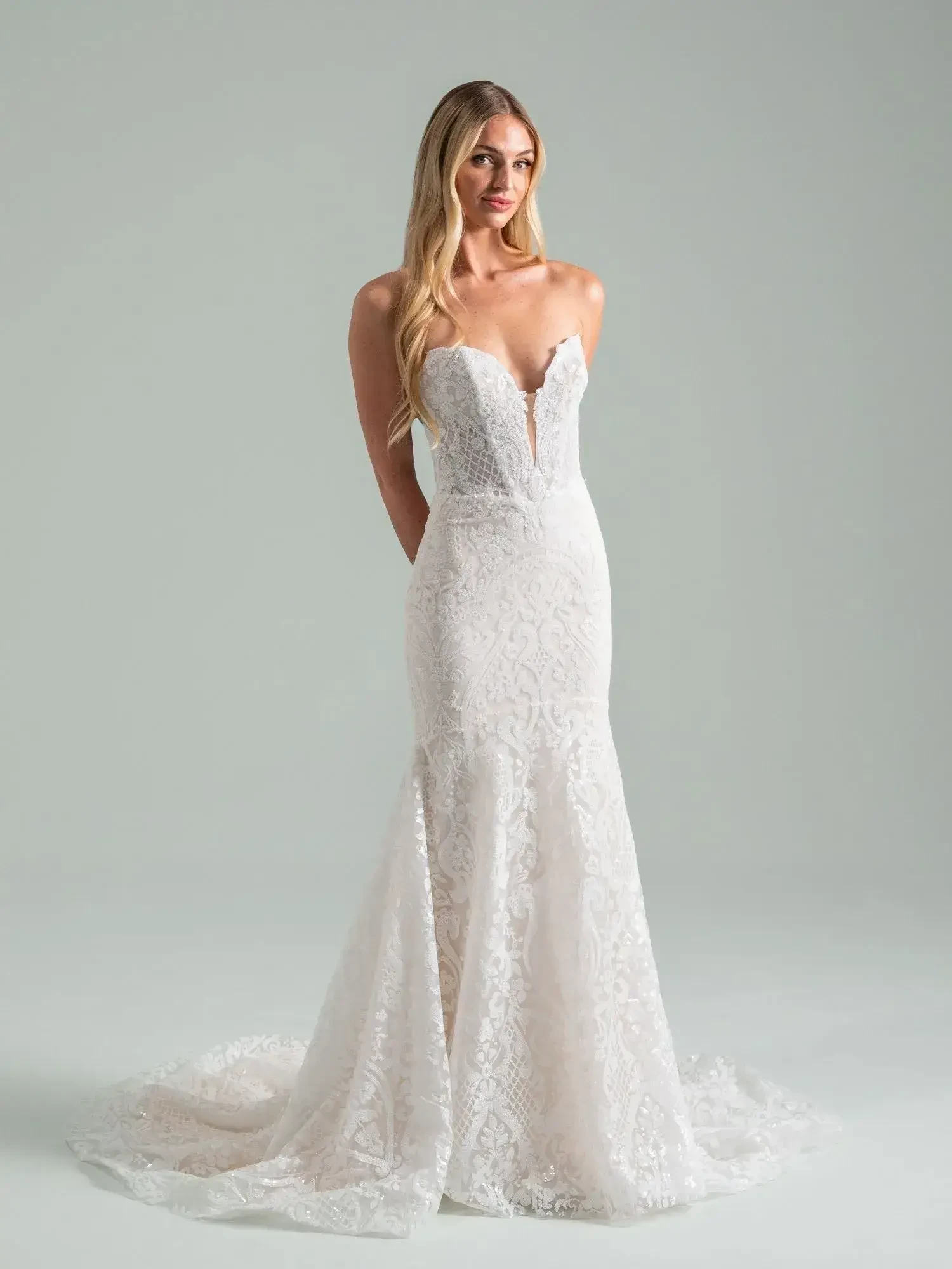 A woman in a strapless, form-fitting lace wedding gown with a sweetheart neckline and long train stands against a plain backdrop, conveying elegance and poise.