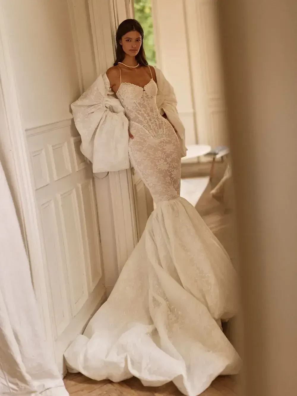 A model wearing a white mermaid-style gown with intricate lace details, standing in a softly lit room with light-colored walls and wooden flooring.
