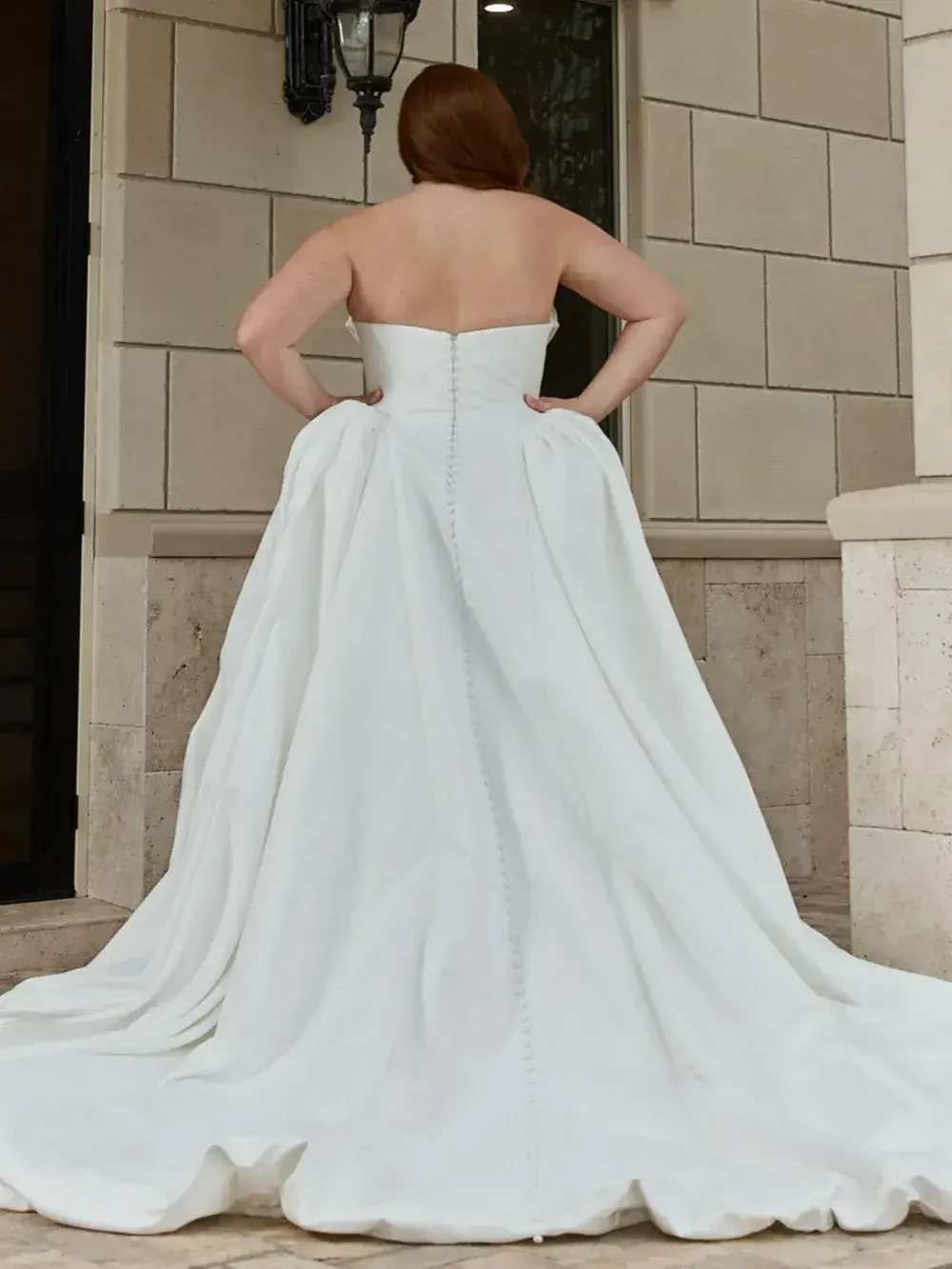 A person standing with their back towards the camera, wearing a strapless white wedding gown with a flowing train.