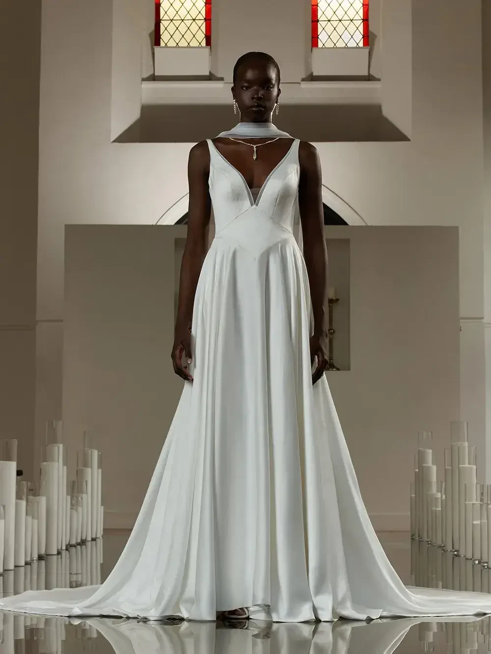 A model wearing an elegant white wedding dress stands in a minimalist indoor setting with tall candles.