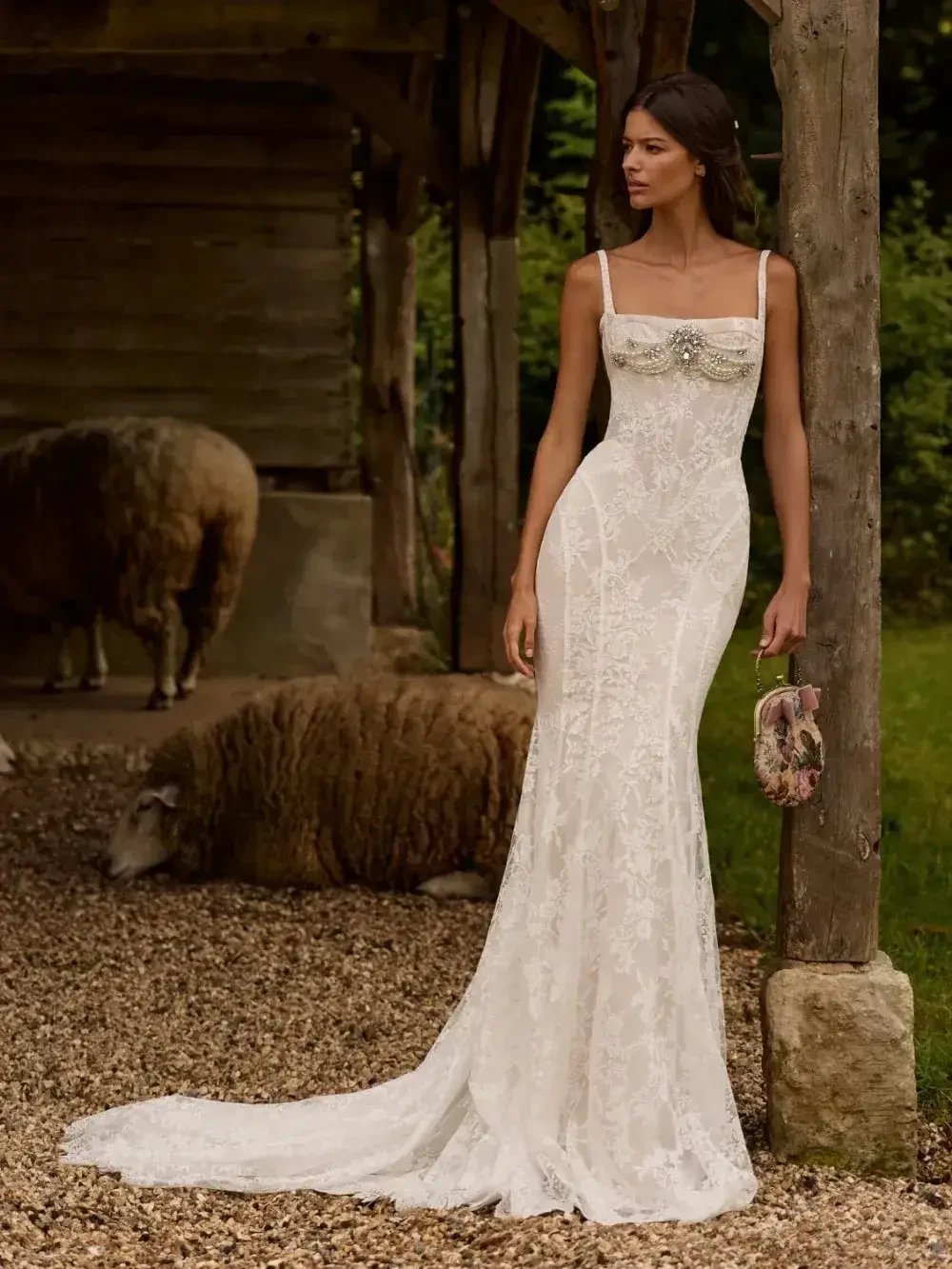 A model wearing a fitted white wedding dress with lace details, standing outdoors near a wooden structure and sheep in the background.