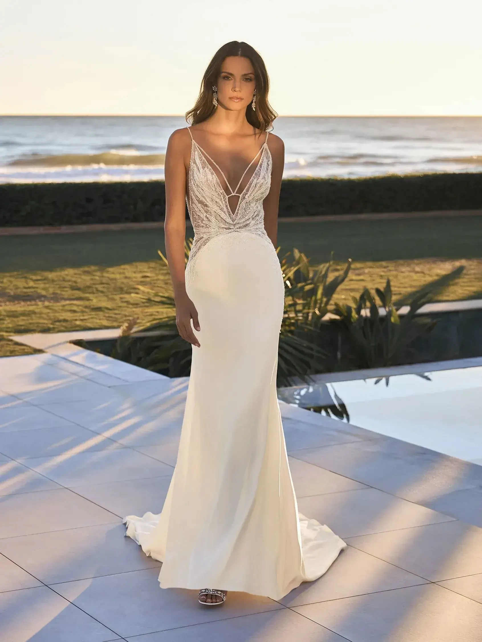 A model wearing a sleek, white gown with intricate bead detailing and a form-fitting silhouette, standing by the ocean during sunset.