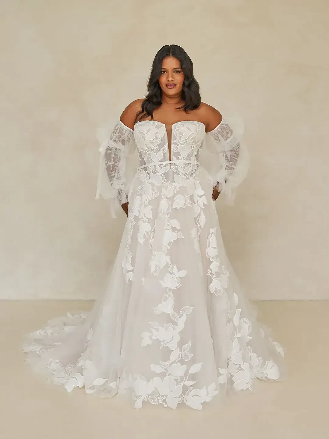 A woman wearing a wedding dress with floral lace detailing and sheer sleeves, posing against a neutral background.