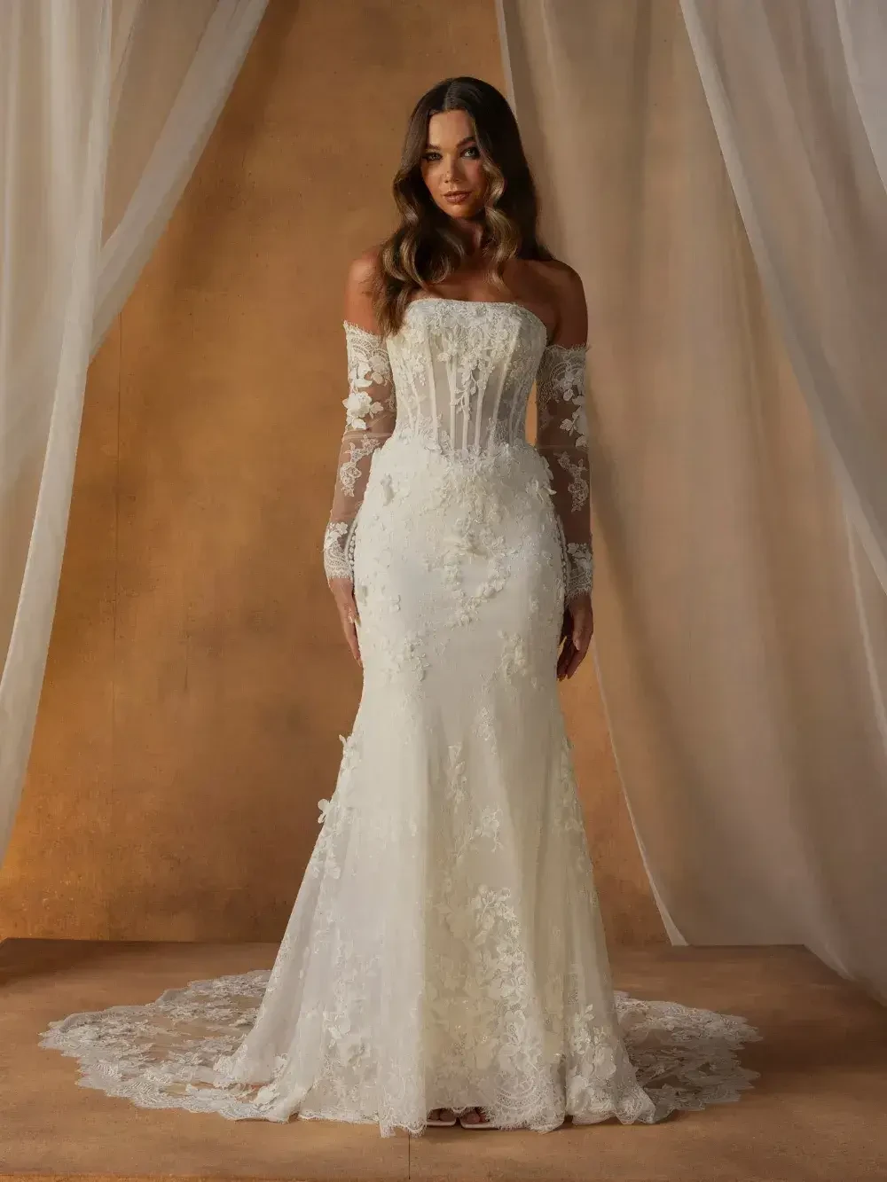 A woman is wearing a white wedding gown with detailed lace and off-the-shoulder sleeves, standing against a soft, neutral backdrop.