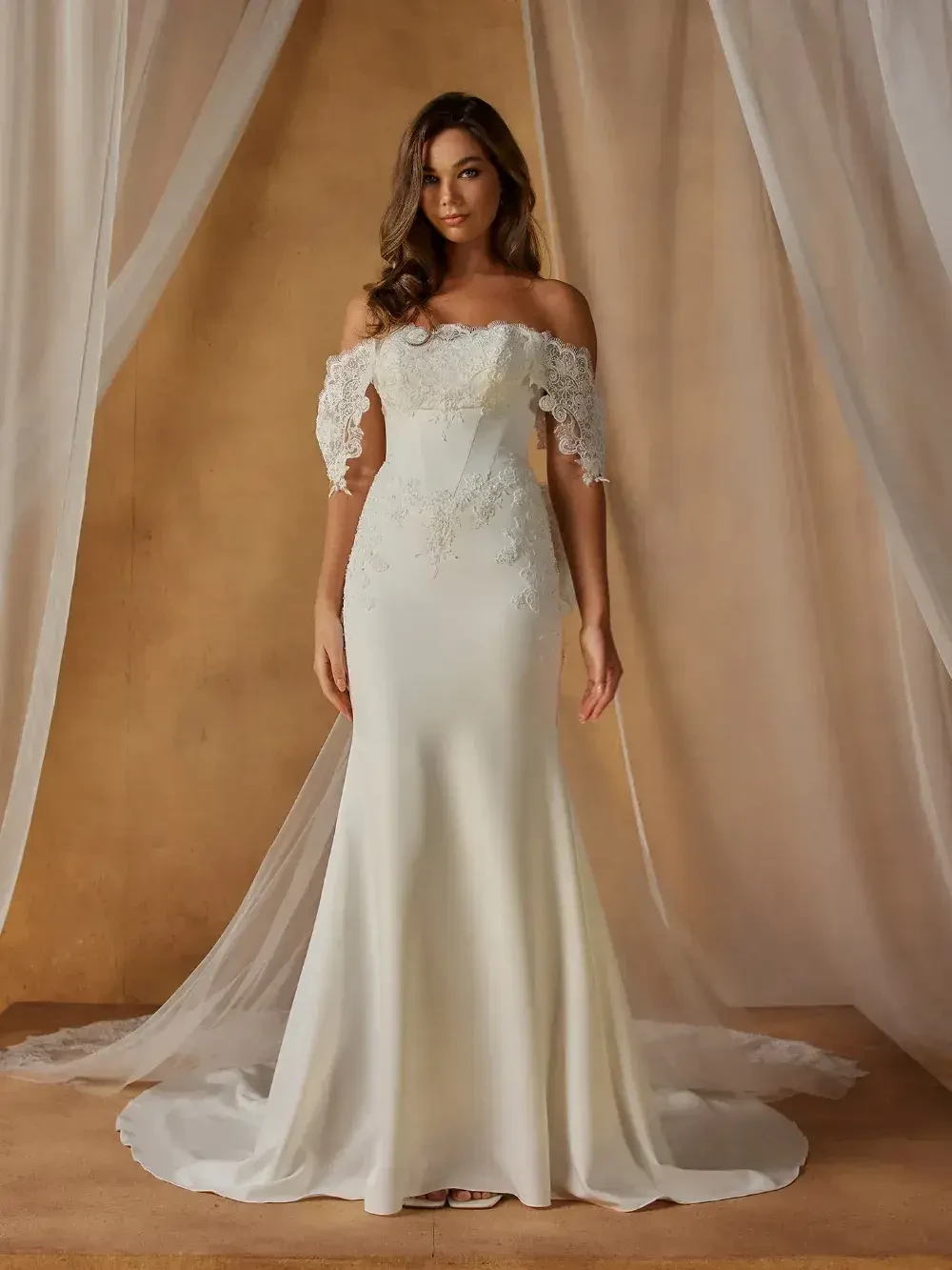 A model wearing an elegant off-shoulder wedding gown with lace details, standing in front of a draped backdrop.