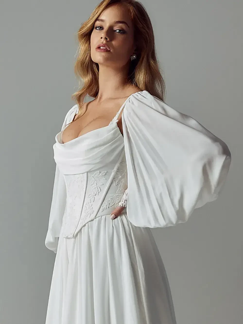 A woman wearing a white dress with long, flowing sleeves and a fitted bodice, posing against a neutral background.