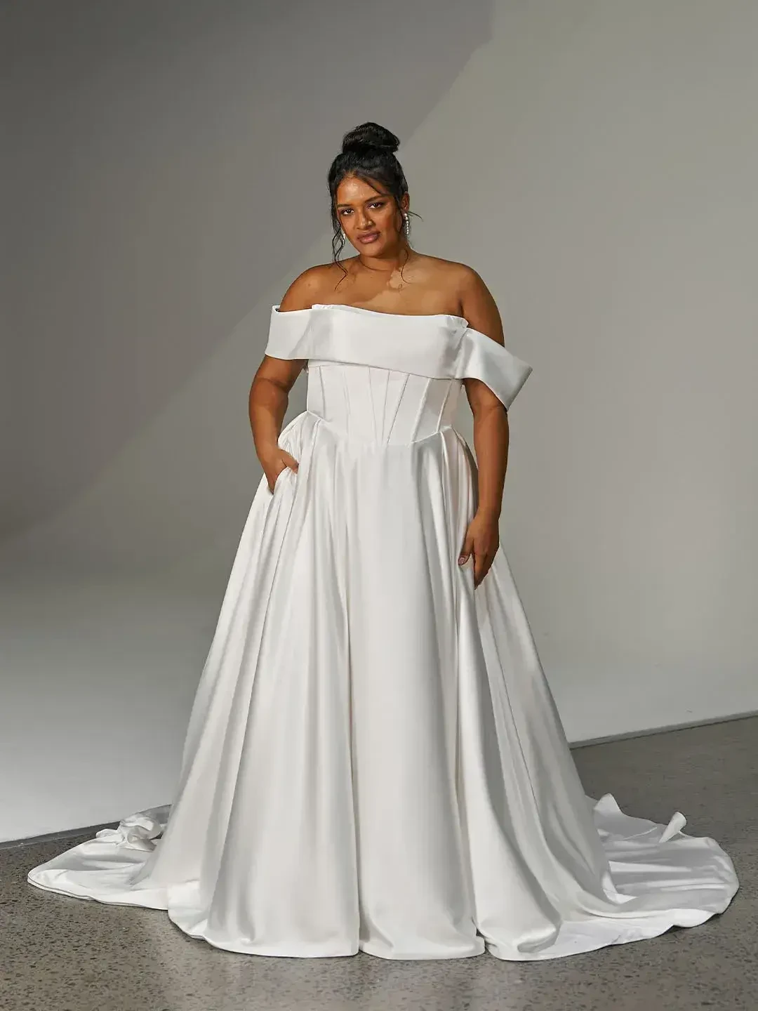 A woman in a white off-the-shoulder gown with pockets, standing against a neutral background.