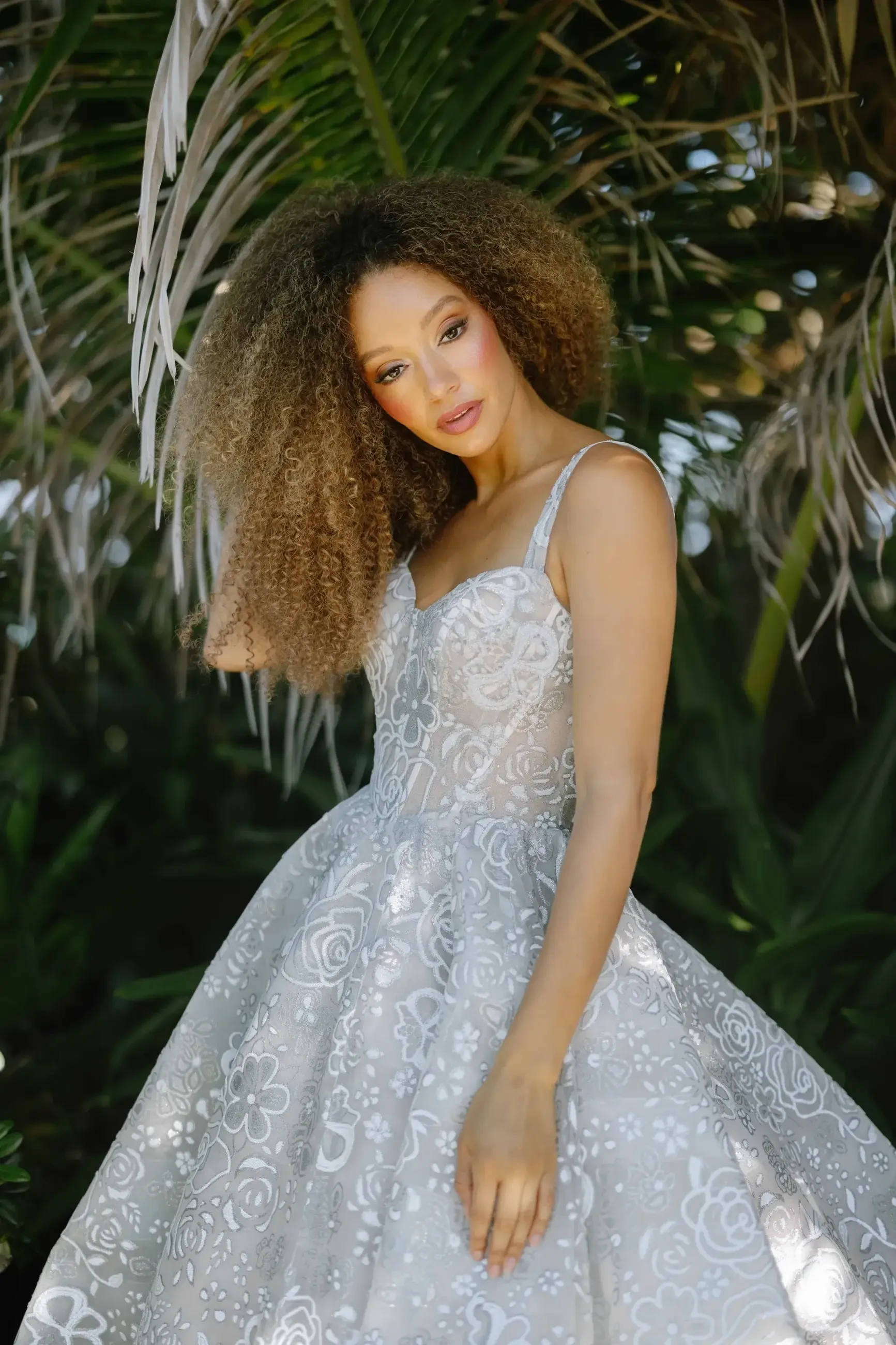 A woman with curly hair wearing a silver dress with floral patterns stands in a lush, green environment.