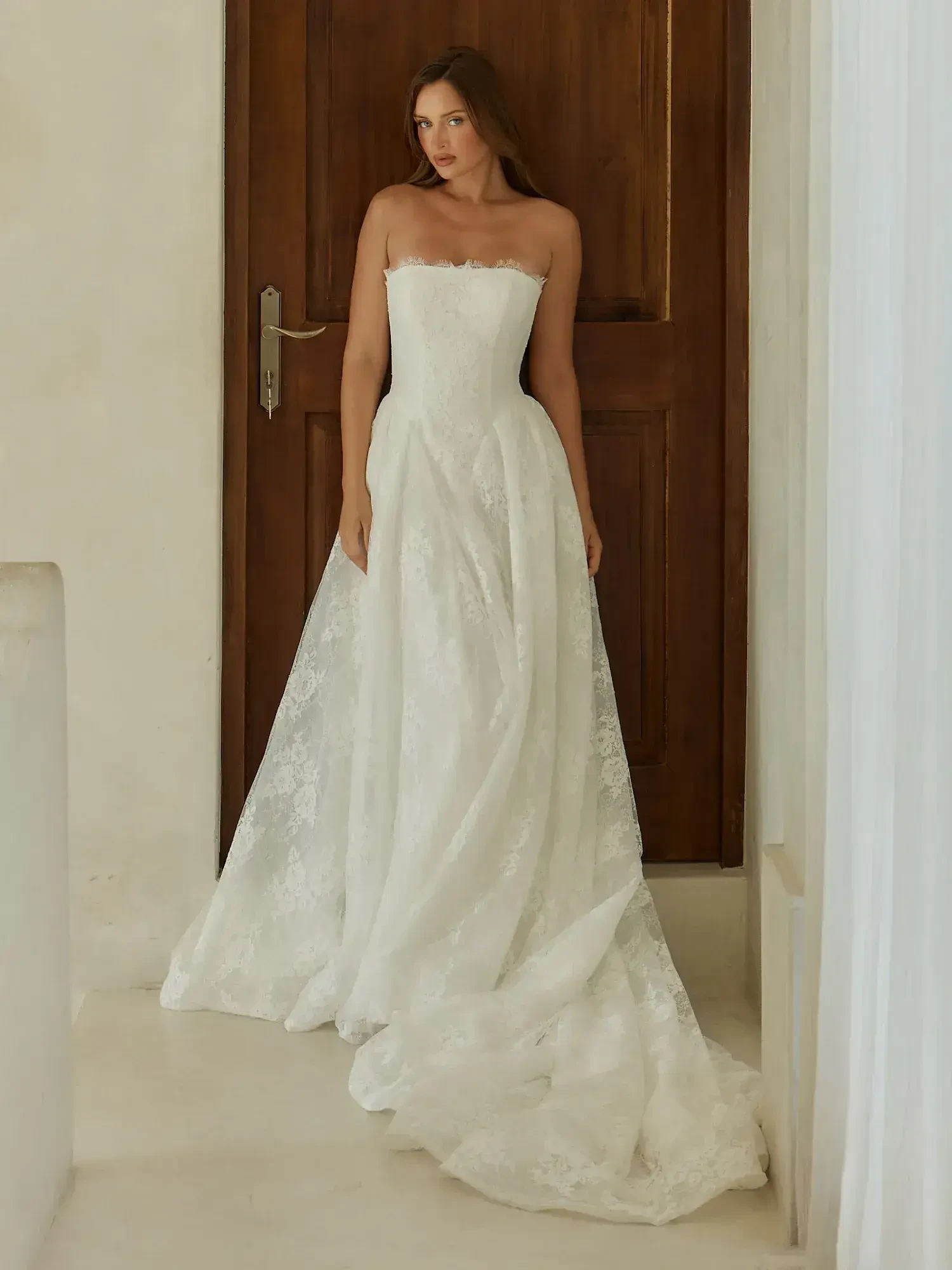 A woman in a strapless white wedding dress stands gracefully in front of a wooden door.